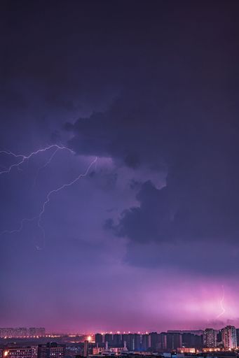 A bright electric bolt of lightning strikes the sky above a modern cityscape, creating an impressive light show in the night sky