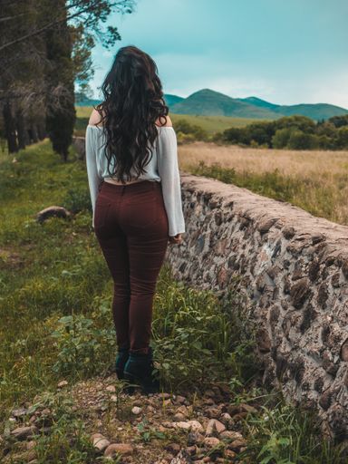 A beautiful shot of a girl with long wavy hair from behind wearing a white shirt and maroon pants in a farm