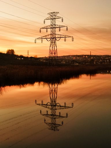 A vertical shot of a transmission tower reflecting on a small pond during a golden sunset