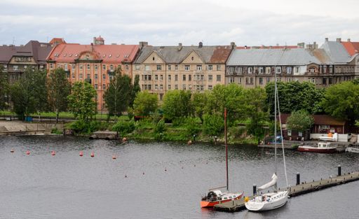 A serene, isolated boat moored in the middle of a small body of water in the foreground, with buildings in the background