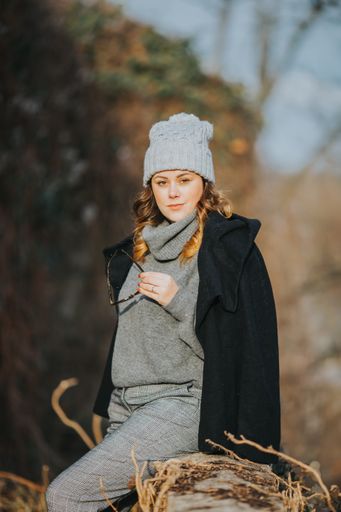 A vertical shot of a young lady wearing a gray turtleneck sweater, trousers, a hat with a black coat