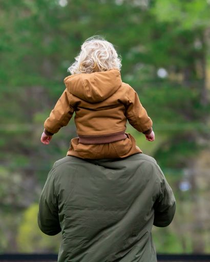 A vertical rear of a young blonde boy sitting on his father's shoulders trees blurred background