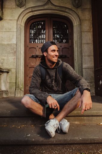 A vertical shot of a young South American man sitting against an old door with a backpack