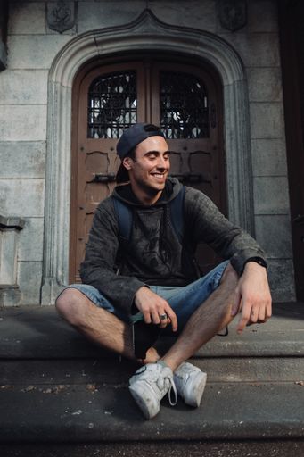 A vertical shot of a young South American man sitting against an old door with a backpack