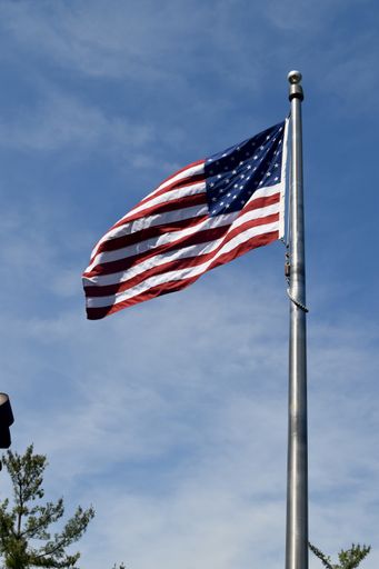 A low angle view of the American flag near trees under a blue cloudy sky and sunlight
