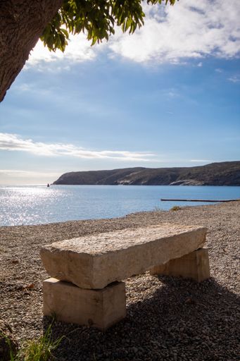 Lonely stone bench looking towards the endless sea with the green mountain and under a sunny blue sky.