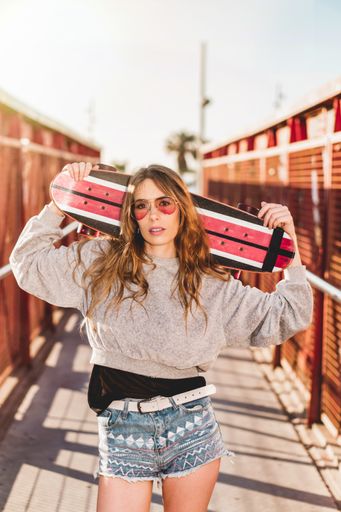 A vertical shot of a cool Caucasian woman wearing sunglasses and holding her skateboard on the bridge
