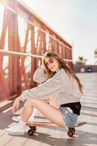 A vertical shot of a cool Caucasian blonde woman wearing sunglasses sitting on a skateboard