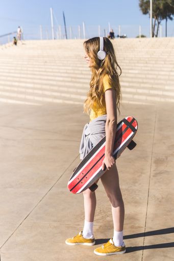 A vertical shot of a young Caucasian woman wearing headphones and holding a skateboard during a sunny day