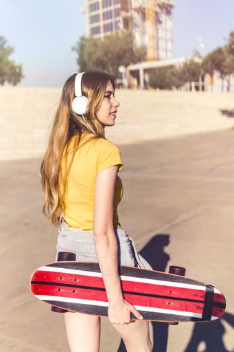 A vertical shot of a young Caucasian woman wearing headphones and holding a skateboard during a sunny day