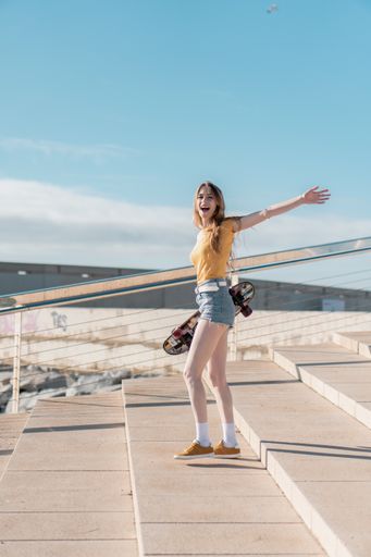 A vertical shot of a Caucasian woman holding a skateboard and happily smiling during a sunny day