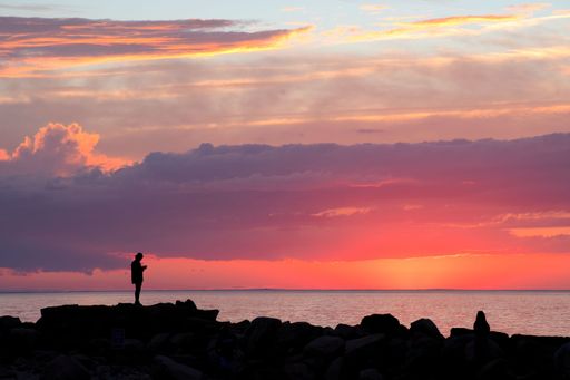 Dark outlines of two unrecognizable people on breakwater rocks at Cape Cod beach viewing colorful sunset just as sun drops below the horizon.