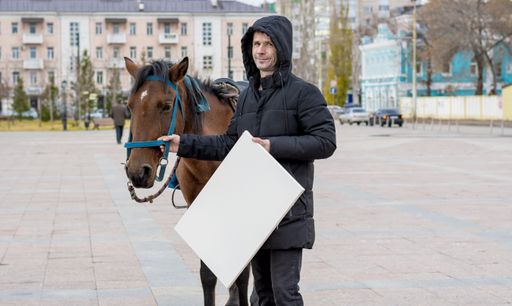 young man holds in one hand blank white canvas for drawing, and with the other hand he holds horse in knots against the background of a cityscape