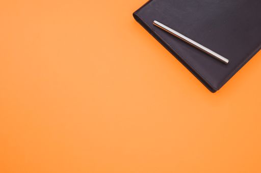 A top view closeup of a black notebook with a yellow bookmark and a pen isolated on an orange background