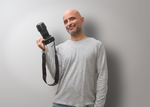 A Hispanic bald man smiling and holding his camera on a gray background