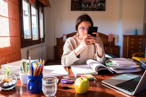 A young female student sitting on her desk in the bedroom using a phone, the concept of studying at home