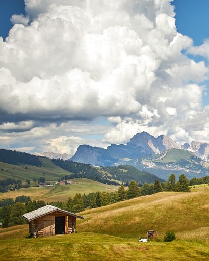 A vertical shot of a wooden cabin in a green land under white clouds with the beautiful mountains in the background
