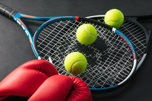 A high angle shot of tennis rackets, tennis ball, and boxing gloves on a black surface