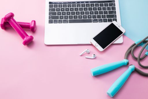 A high angle shot of a laptop and phone, a jumping rope, and dumbbells on a pink surface