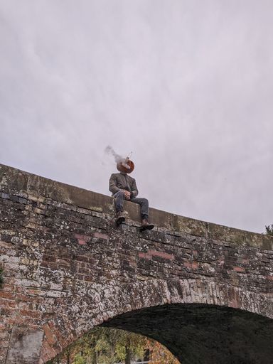 Vertical shot of a person with a pumpkin on his head and smoke coming out while sitting on a bridge