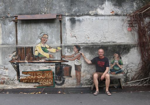 With a painted girl on a bench sitting real Caucasian man in 3D street art mural in Georgetown, Malaysia, February 2, 2020. Boy buying from old lady.