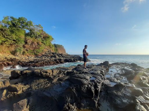 Young wet Asian man in profile view standing daydreaming on a rock plateau with a bag of chips at Wediombo Beach, Java, Indonesia