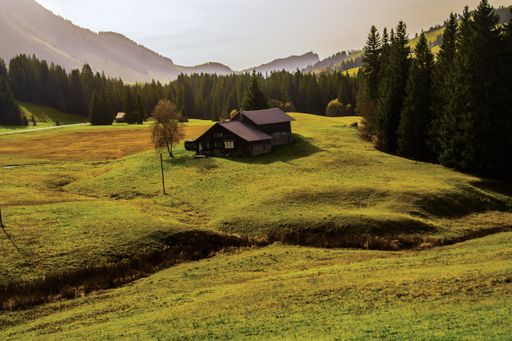 A single-story rural house situated on a lush green hill overlooking a majestic mountain range on a sunny day