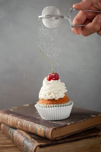 A vertical shot of pouring powdered sugar on a delicious cupcake with cream and cherry on top
