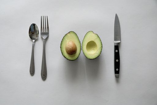 A high angle shot of an Avocado, a knife and spoon and fork on a white surface