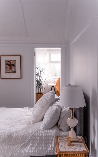 The bedroom with a white bed, a bedside lamp, and a wall picture next to the room with a big green plant