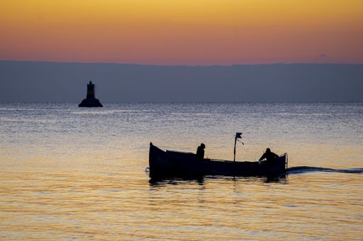 A silhouette of two fishers on a boat fishing at sunset with a lighthouse in the background