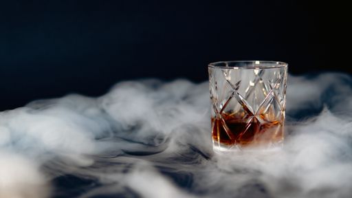 A glass of whiskey on a table covered with smoke against a black background