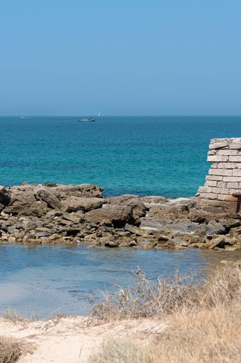 A vertical shot of rocks and wall ruins in the sea under a blue sky and sunlight