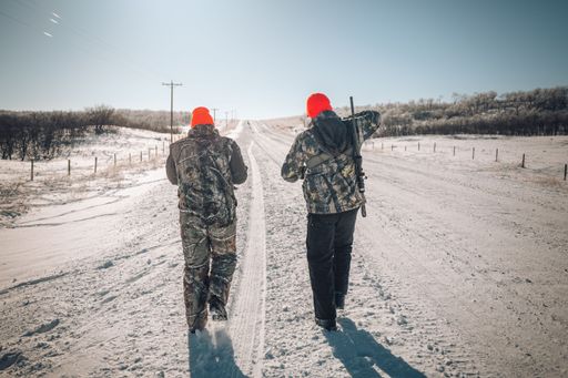 Two male hunters wearing brown caps and heavy winter clothing walking together in a snowy forest,
