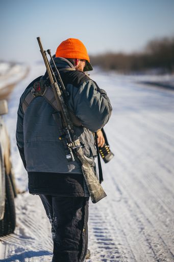 A hunter with a camouflage rifle slung on his back and wearing a bright orange hat walks on a snowy path.