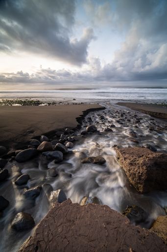 A vertical shot of a stream with long exposure surrounded by rocks under a cloudy sky