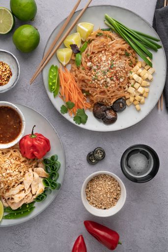 A vertical shot of servings of delicious noodles with salads, lime, greens, sauce, and crumbs