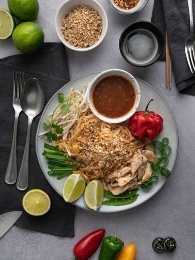 A vertical shot of a serving of delicious noodles with salad, lime, greens, sauce, and peppers