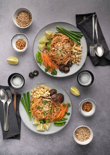 A vertical shot of servings of delicious noodles with salad, lime, greens, carrot, and crumbs