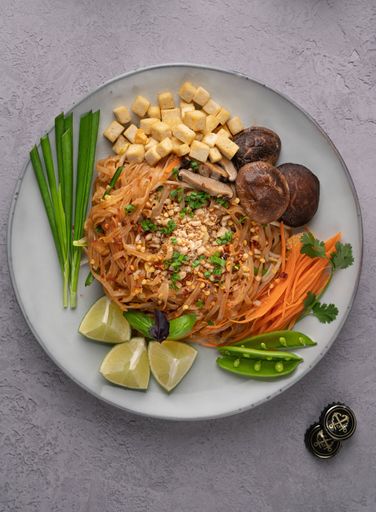 A vertical shot of servings of delicious noodles with salad, lime, greens, carrot, and crumbs