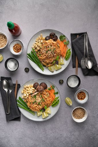 A vertical shot of servings of delicious noodles with salad, lime, greens, carrot, and crumbs