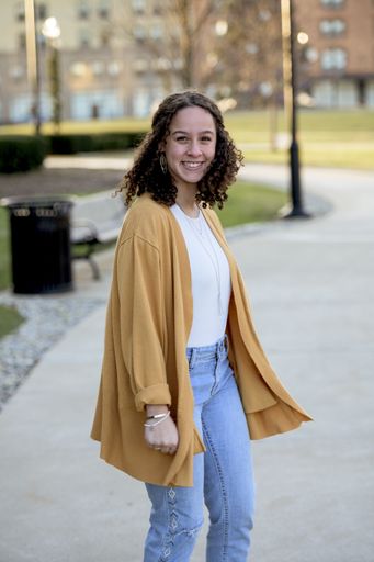 A woman with curly hair wearing a yellow jacket in a park under sunlight with a blurry background