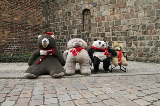 Four stuffed bear toys on chairs outdoors against an old stone bu