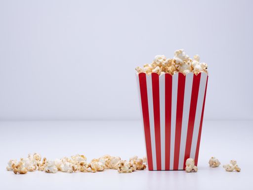 A paper cup of popcorn and some popcorn scattered around on a white surface