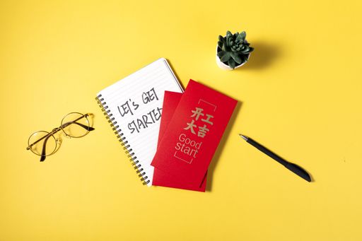 The flat lay of glasses, notepad, Chinese red envelope, pen, and flower pot on the yellow background, motivating inscriptions for starting a business