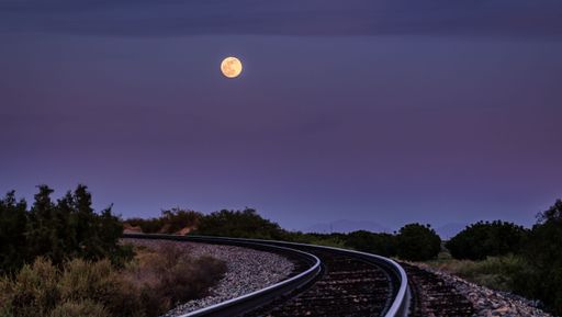 The full moon in purplish blue sunrise sky over winding train tracks in the countryside