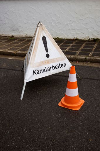 An isolated triangular warning sign with an exclamation point symbol in the center, set near an orange traffic cones