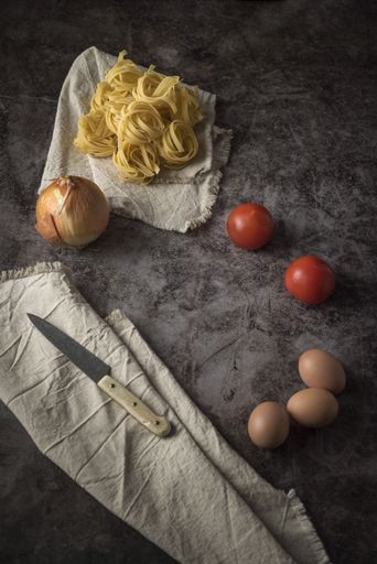 An overhead shot of fresh onions, eggs, tomatoes and raw pasta with knife and table cloth on a stone table
