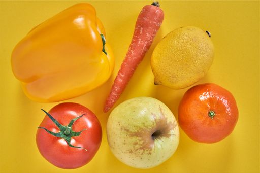 A closeup shot of vegetables and fruits: pepper, lemon, apple, tangerine, tomato, carrot, yellow background