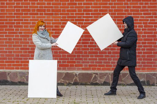 Young man and woman standing against a background of a red brick wall holding three white canvases in front of them to paint